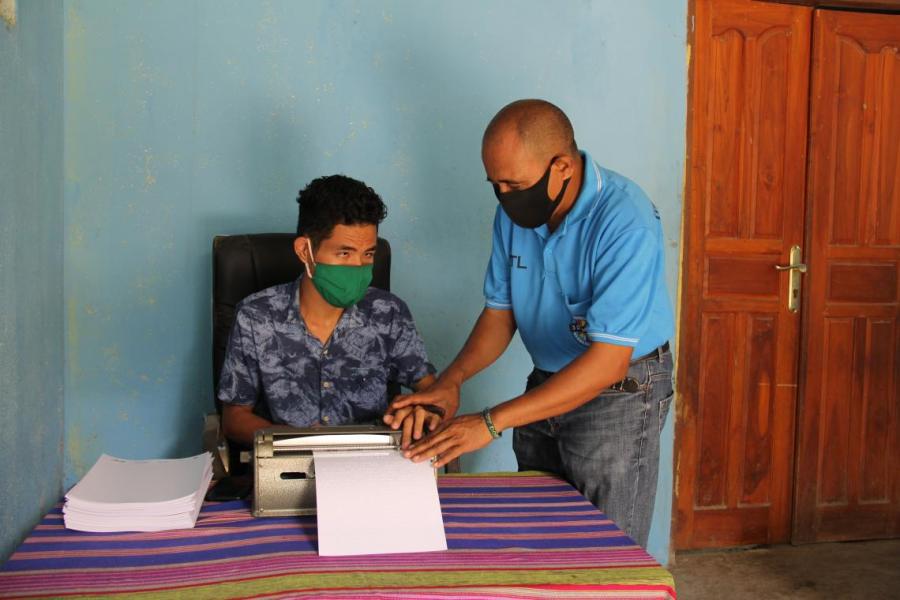 Domingos and his assistant João work on a braille machine to create documents containing COVID-19 prevention messaging for people with a vision impairment.